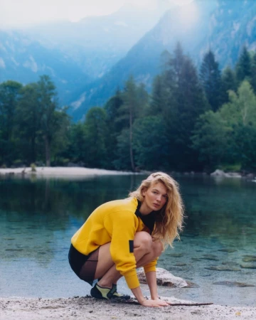 Model Caitlin Soetendal crouches by a clear alpine lake wearing a yellow sweatshirt and dark shorts against a mountainous landscape backdrop.