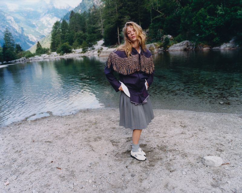 Caitlin Soetendal standing by an alpine lake wearing a fringed jacket and pleated skirt for Pop Magazine.