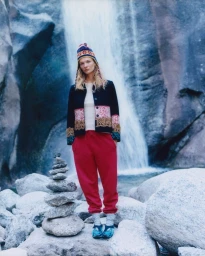 Model Caitlin Soetendal wearing a patterned jacket, red trousers and a beanie hat standing in front of a waterfall