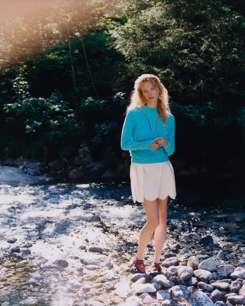 Model wearing a blue sweater and white skirt standing on a rocky riverbank in a forest for Pop Magazine