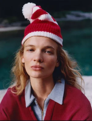 Model Caitlin Soetendal wearing a red and white striped beanie and a maroon zip-up top against a dark water background.