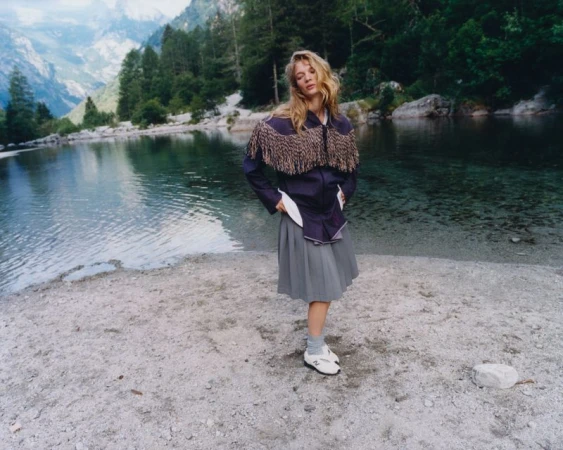 Caitlin Soetendal standing by an alpine lake wearing a fringed jacket and pleated skirt for Pop Magazine.