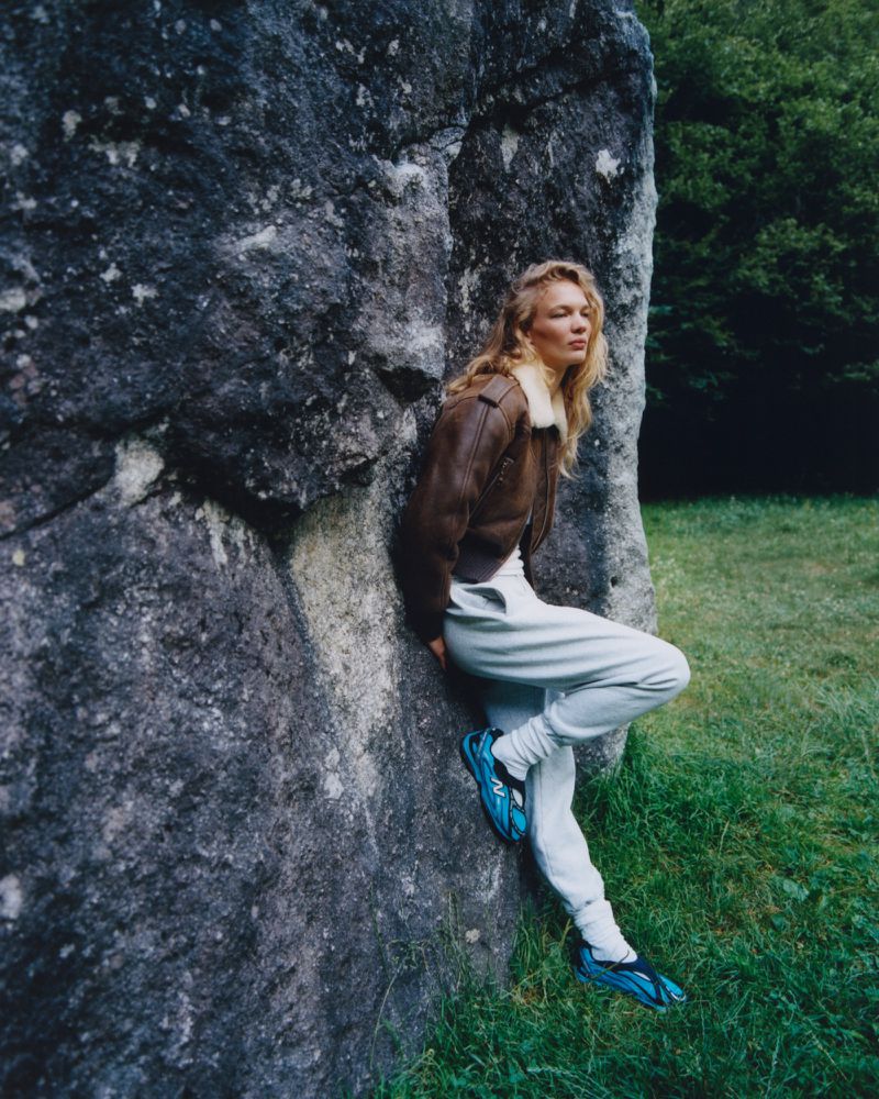 Model leaning against a large rock face wearing a brown leather jacket and sweatpants in an outdoor setting