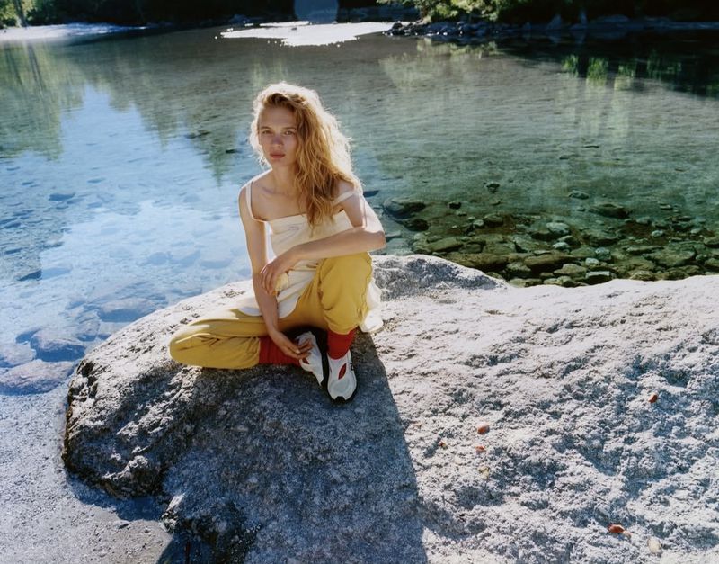 Model Caitlin Soetendal sitting on a large rock by clear river water wearing yellow trousers and white sneakers for Pop Magazine.