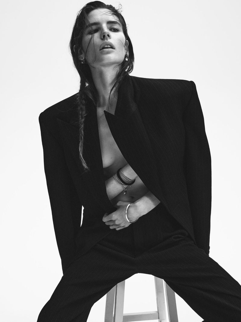 Model with braided damp hair wearing a structured dark blazer and trousers seated on a white stool against a light background