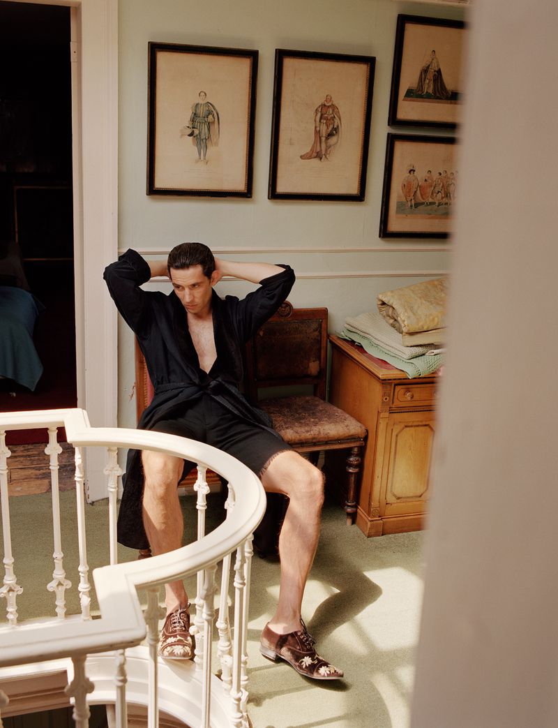 Actor Josh O Connor in a dark robe seated near a stairway for AnOther Magazine by Alasdair McLellan