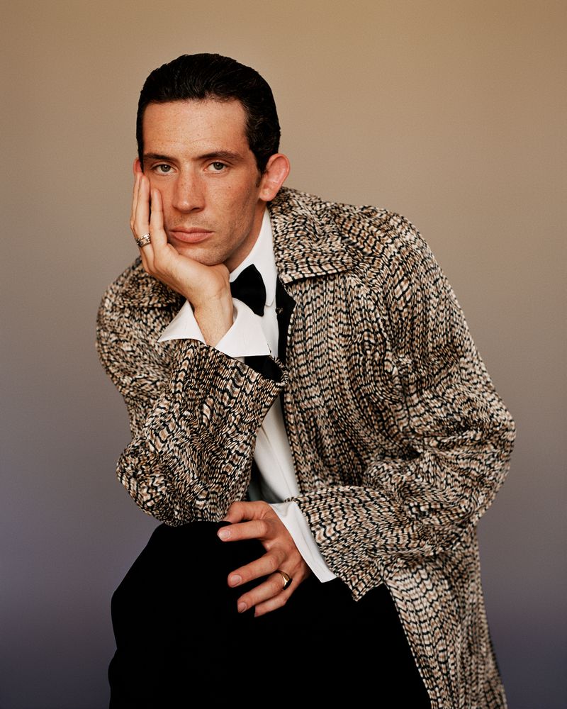 Josh O'Connor in a patterned tweed jacket and white shirt with a black bow tie, resting his face in his hand against a neutral background.