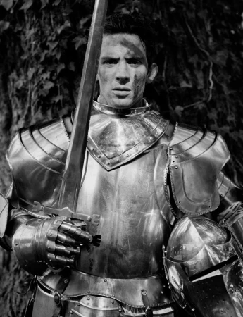 Man wearing polished metal armor holding a vertical sword outdoors in black and white