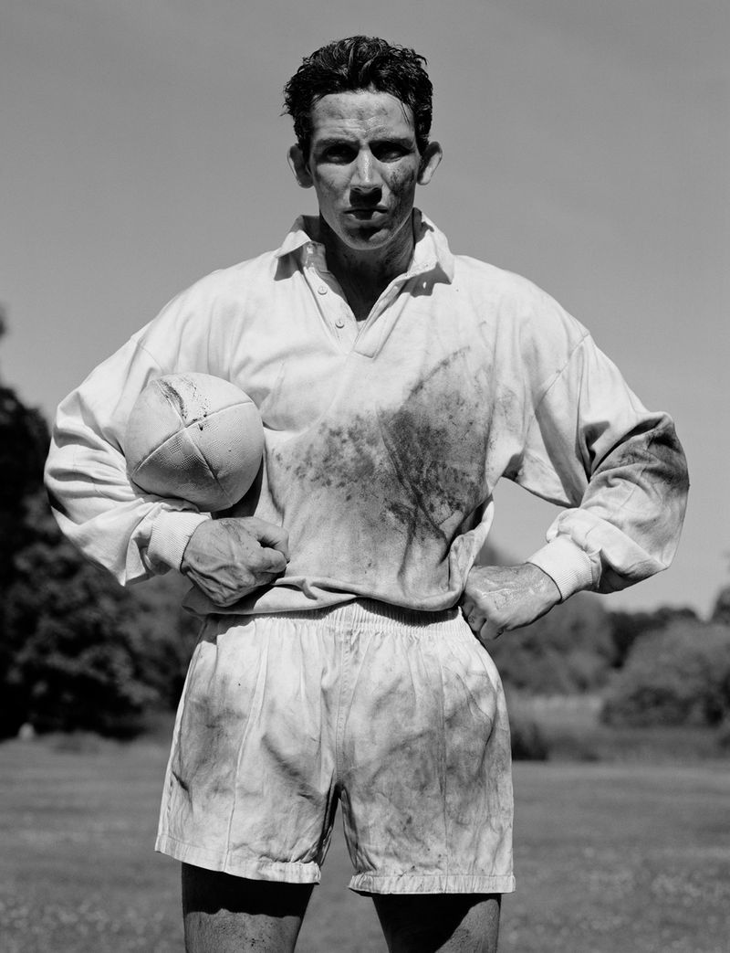 Man wearing a smudged white rugby jersey holding a rugby ball outdoors against a clear sky