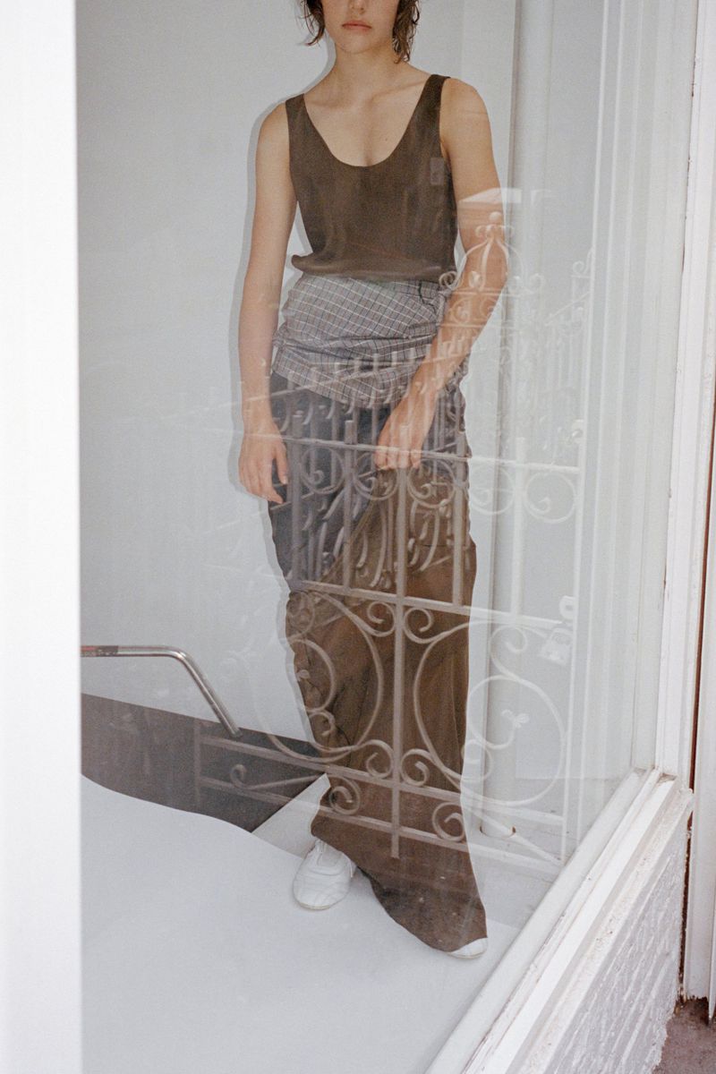 Model wearing a dark sleeveless top and long skirt behind a glass door with white ornate iron security bars.