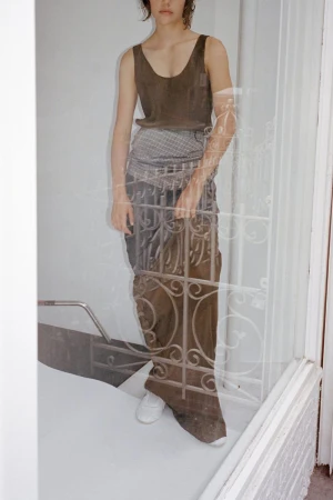 Model wearing a dark sleeveless top and long skirt behind a glass door with white ornate iron security bars.