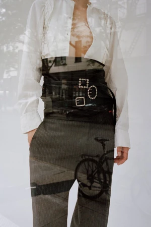 Model wearing a white button-down shirt, a wide buckled black waist belt, and pinstripe trousers with a double exposure reflection.