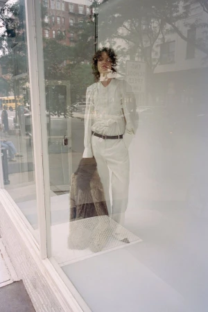 Model standing behind a glass storefront wearing light-coloured trousers and holding a furry jacket for CDLM.