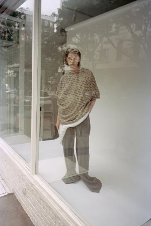Model standing behind a storefront window wearing an patterned oversized top and grey trousers in CDLM lookbook