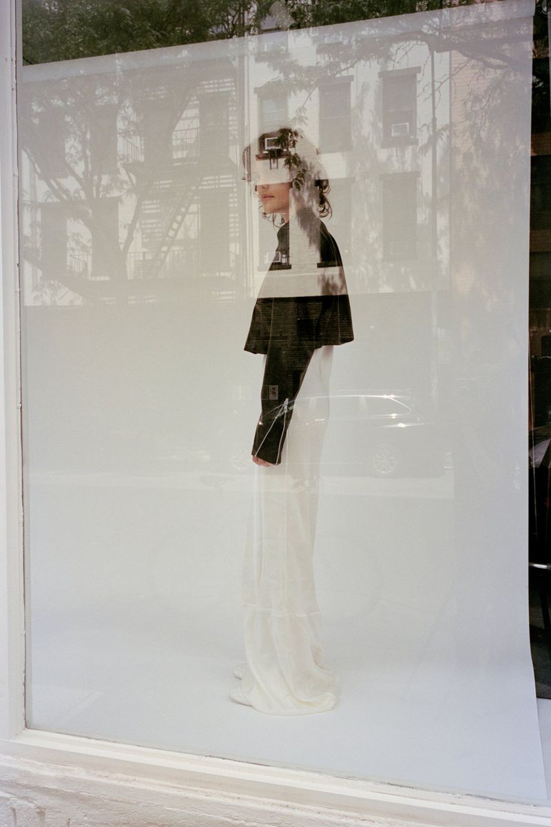 Model standing against a white seamless backdrop viewed through a glass window reflecting a street scene.