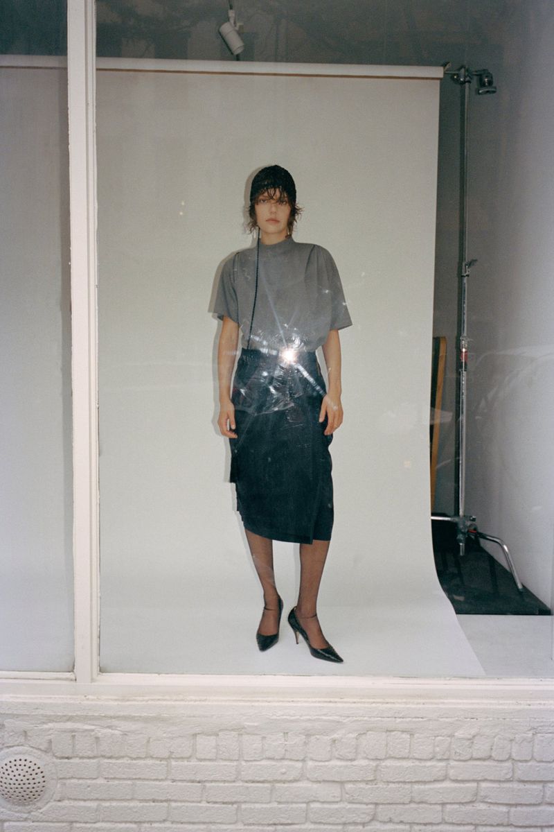 Model wearing a grey t-shirt and dark skirt standing in front of a white seamless backdrop viewed through a window.