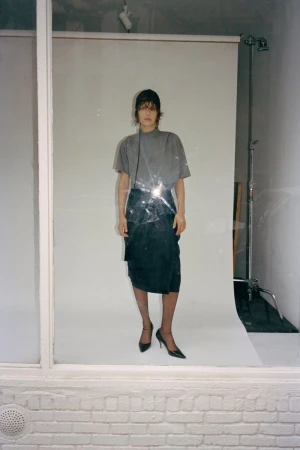 Model wearing a grey t-shirt and dark skirt standing in front of a white seamless backdrop viewed through a window.