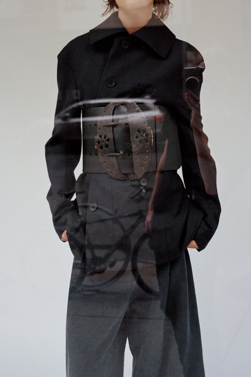 Model wearing a black coat with a wide decorative leather belt against a white backdrop
