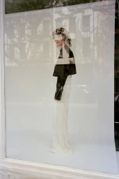 Model standing against a white seamless backdrop viewed through a glass window reflecting a street scene.