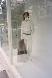 Model standing behind a glass storefront wearing light-coloured trousers and holding a furry jacket for CDLM.