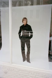 Model wearing a dark jacket and trousers with a wide sculpted belt standing against a seamless white background.