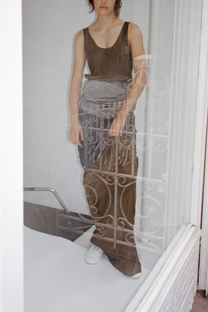 Model wearing a dark sleeveless top and long skirt behind a glass door with white ornate iron security bars.