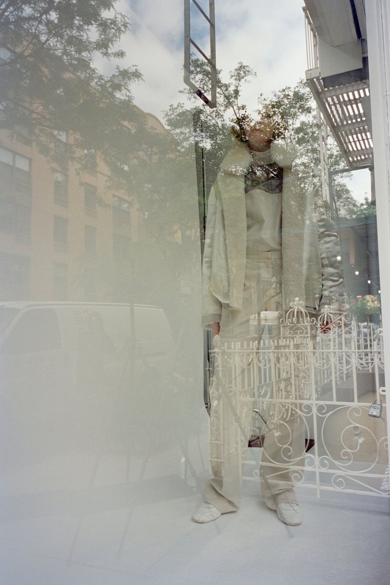 Model standing behind a glass window reflecting a street scene, wearing layered white clothing and a light jacket.