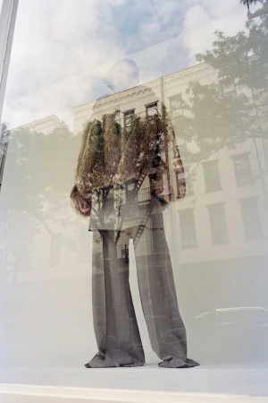 Model wearing tailored grey trousers and structured jacket reflected in a glass window with city buildings