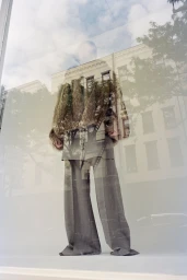 Model wearing tailored grey trousers and structured jacket reflected in a glass window with city buildings