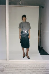 Model wearing a grey t-shirt and dark skirt standing in front of a white seamless backdrop viewed through a window.