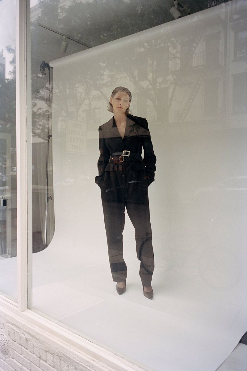 Model standing on a white seamless backdrop viewed through a glass window showing exterior reflections