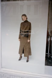 Model standing on a white seamless backdrop wearing a brown turtleneck top and matching skirt with double belts.