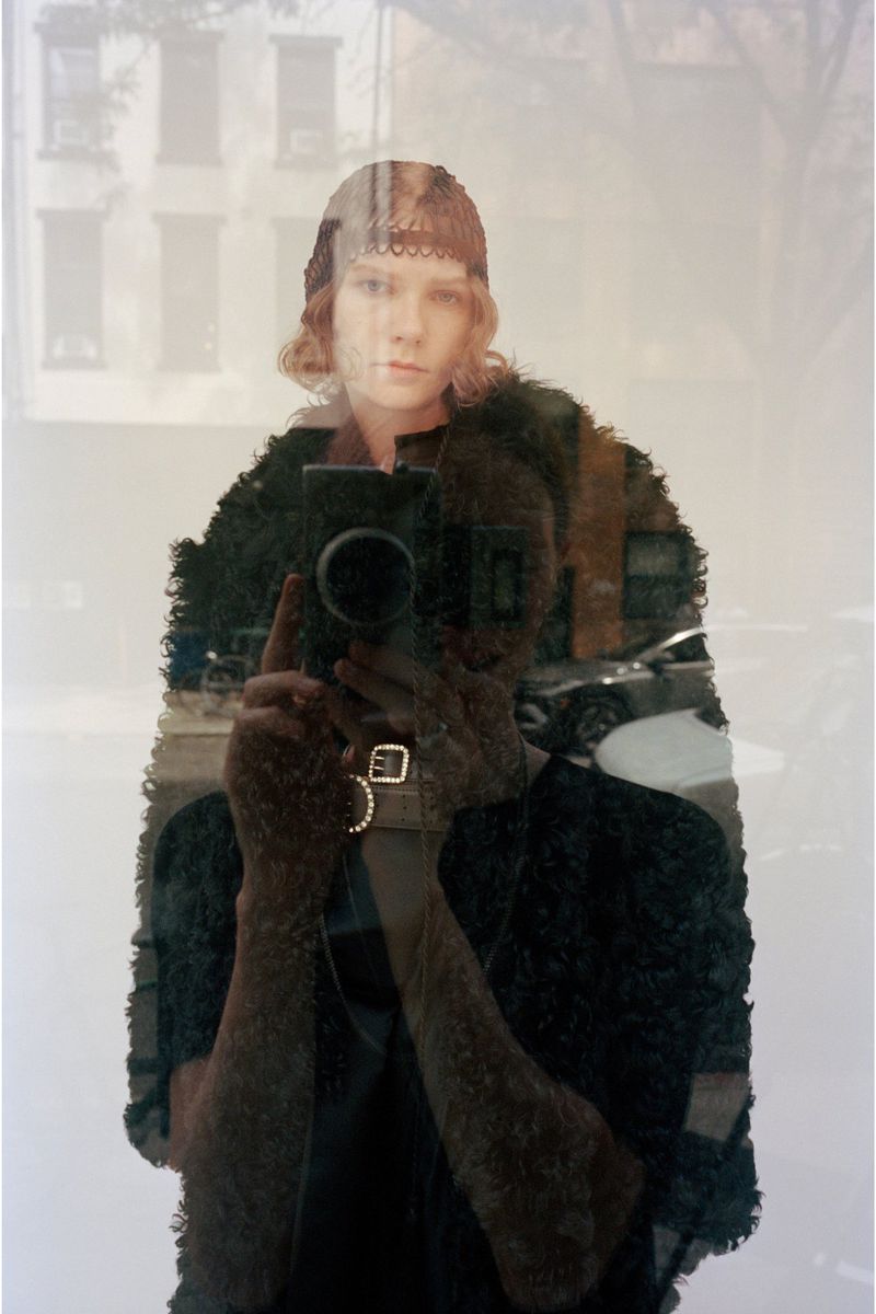 Model in a dark textured garment holding a vintage camera for a mirror reflection shot in a CDLM lookbook.