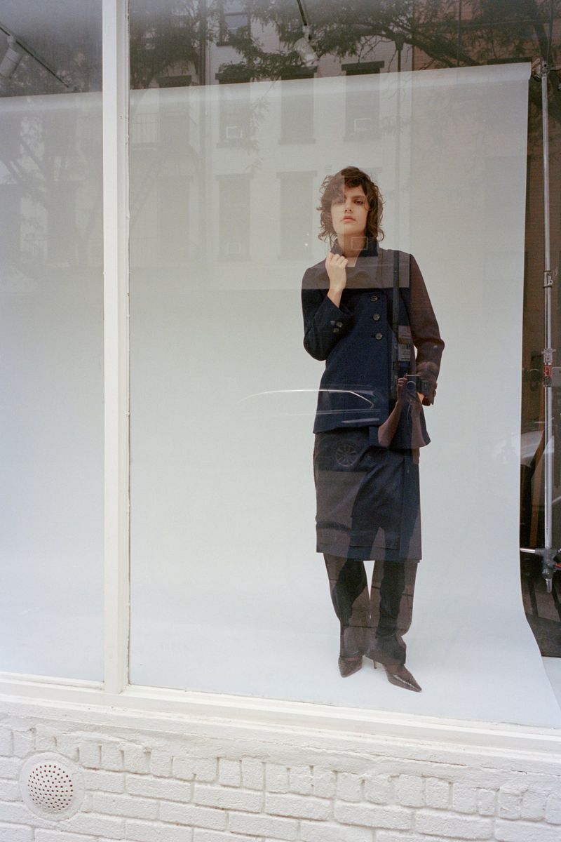Model standing behind a glass window on a white seamless backdrop wearing dark jacket and skirt