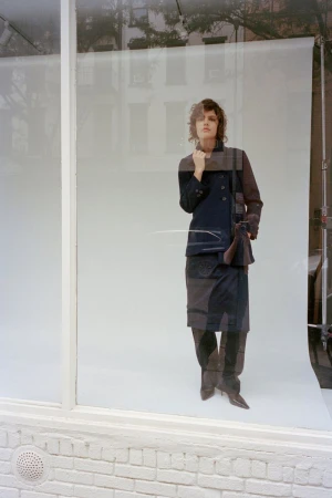 Model standing behind a glass window on a white seamless backdrop wearing dark jacket and skirt