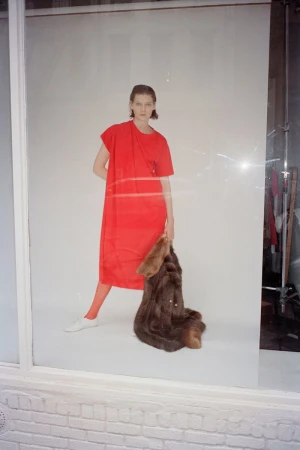 Model wearing a vibrant red dress and holding a brown fur coat against a white studio background
