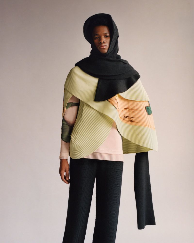 Fashion-forward model showcases a pleated outfit with a unique black head covering against a neutral backdrop in a studio setting.