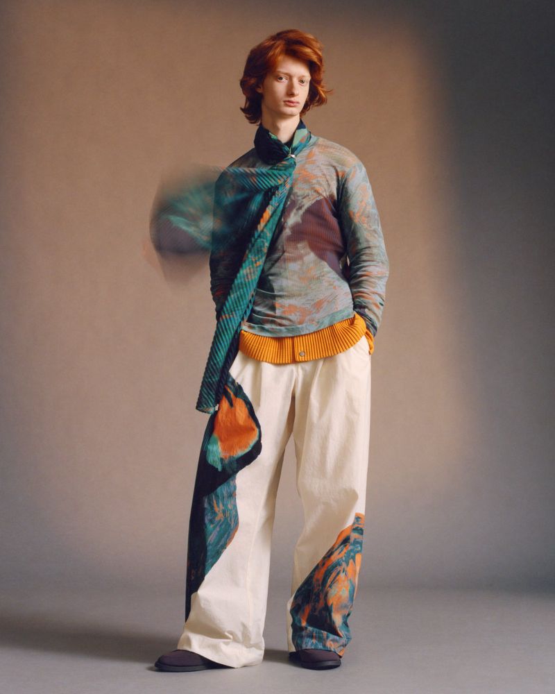 A person stands in a studio with a patterned shirt and pants, featuring a unique scarf and neutral backdrop, ideal for fashion content.
