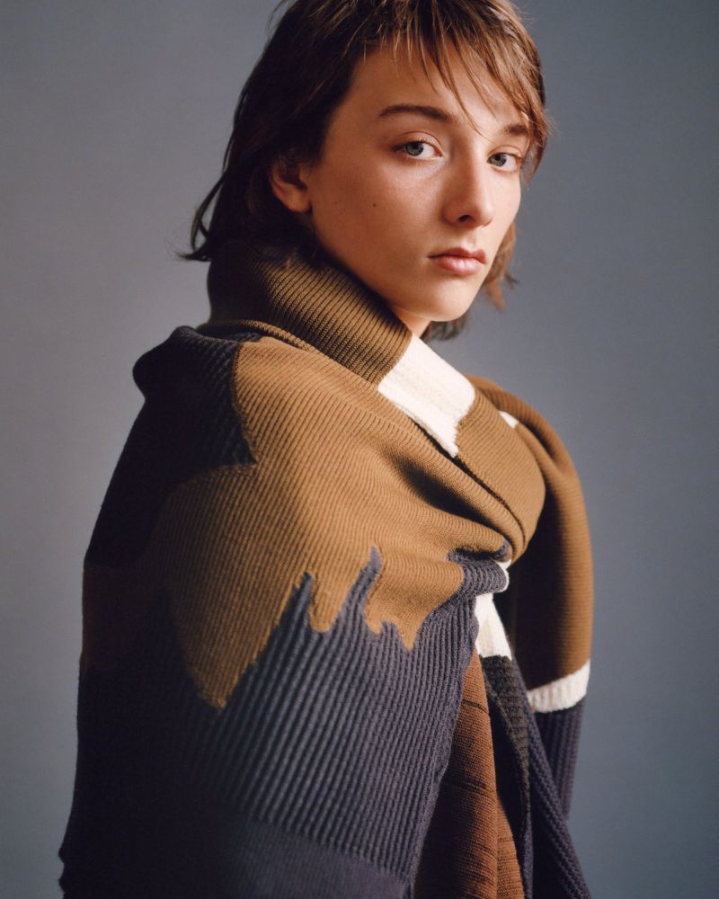 Portrait of a young adult female with short brown hair wrapped in a multi-colored knit scarf against a neutral grey backdrop in a studio.