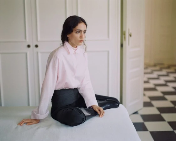 Model seated on a bed wearing a pink shirt and dark trousers in an interior room with panelled doors.