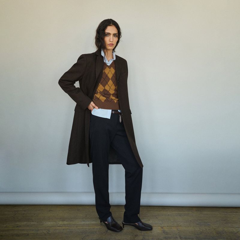 Model wearing a brown coat and argyle sweater over dark trousers against a neutral background
