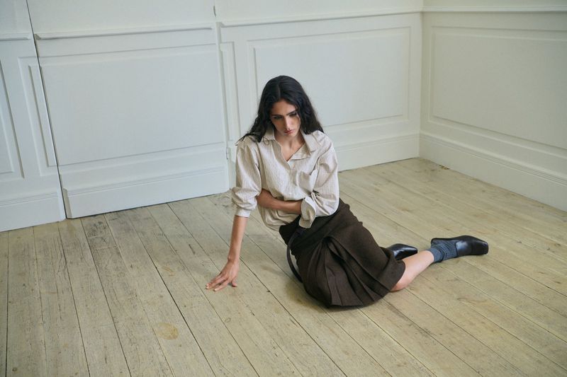 Model sitting on a wood floor wearing a light button-down shirt and dark skirt against white wall panels