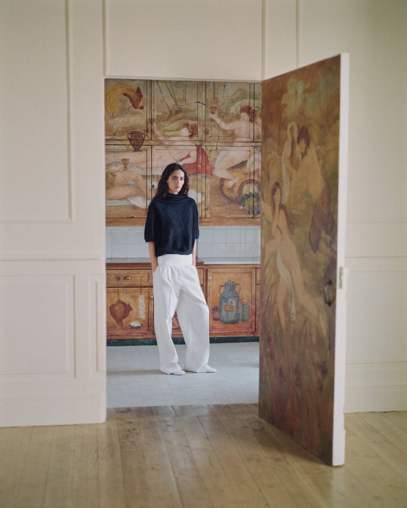 Model standing in a doorway wearing a dark top and white wide-leg trousers before a painted wooden cabinet.