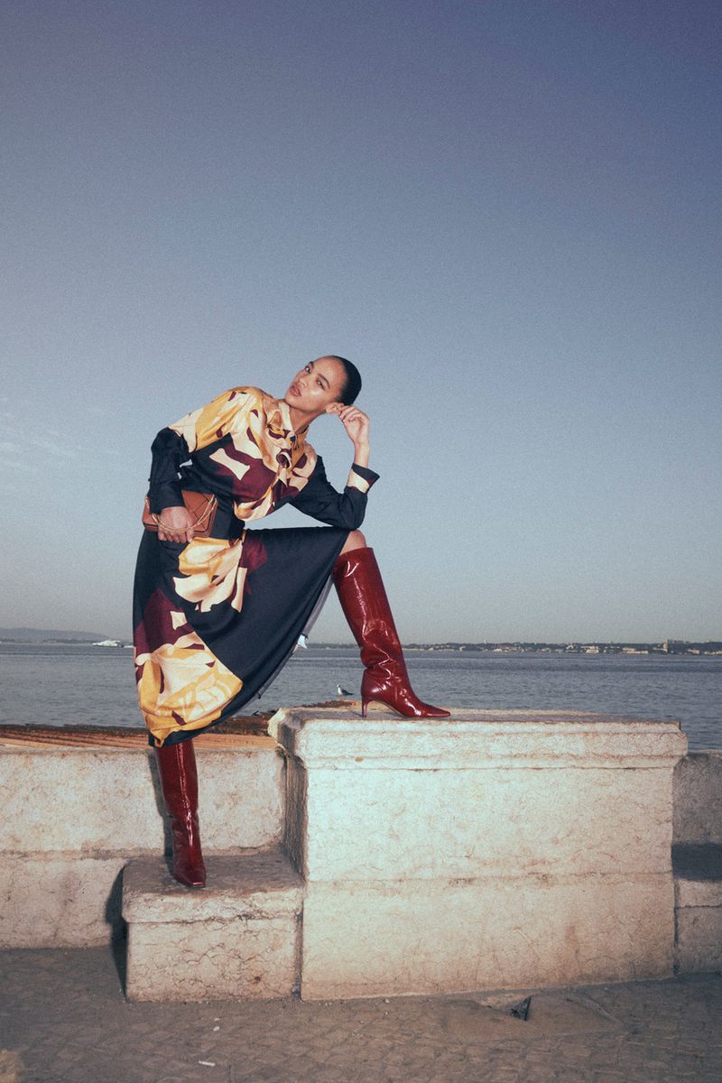 Model posing on a stone wall by the ocean wearing a patterned dress and red boots for Ulla Johnson Resort 2027.