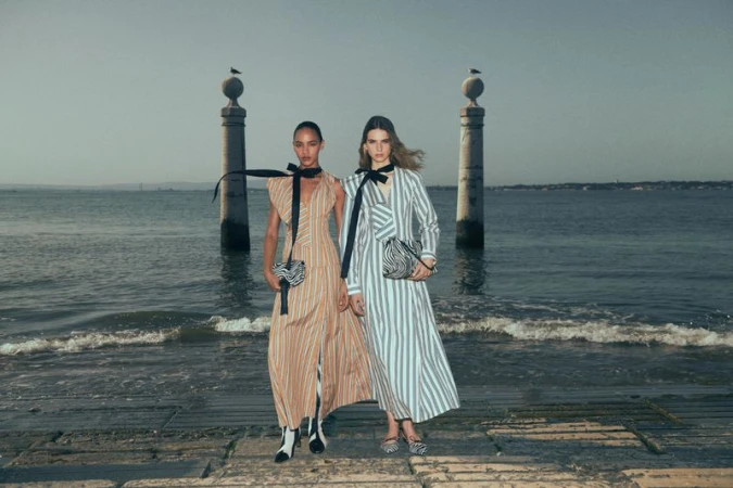 Two female models posing in striped dresses by the water with stone pillars behind them, photographed by Julia Noni for Ulla Johnson Resort 2027.