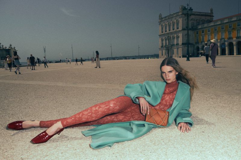 Model reclining in a public square wearing a light blue coat, patterned red outfit, and holding a tan handbag.