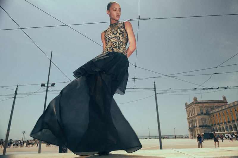 Model wearing a patterned top and flowing dark skirt outdoors against a plaza with overhead power lines and classical architecture.