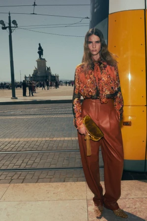 Model in patterned floral blouse and rust trousers standing on an outdoor plaza holding a gold velvet clutch
