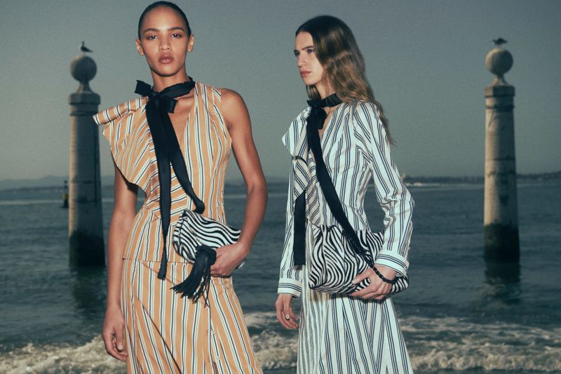 Two female models in striped dresses standing by the water with stone pillars under an overcast sky for Ulla Johnson Resort 2027.