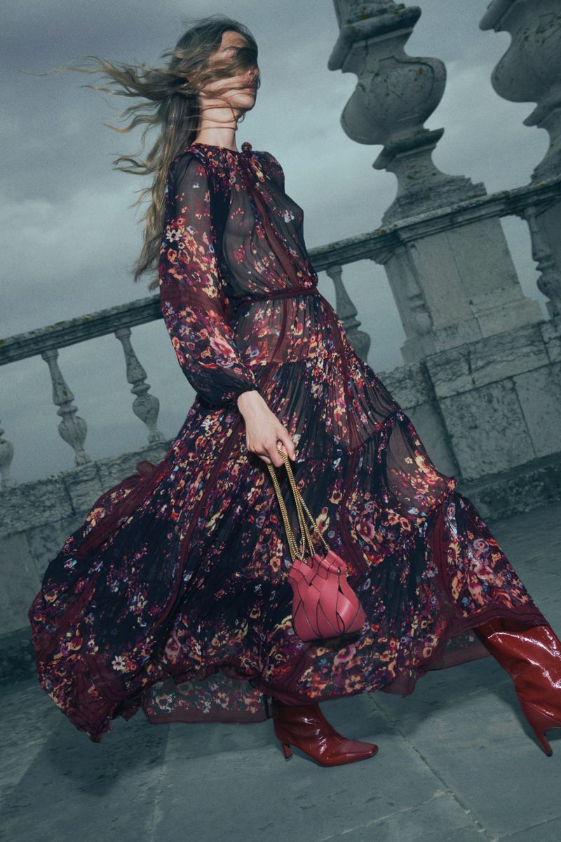 Model in a floral sheer maxi dress and red boots holding a pink leather handbag against a stone balcony